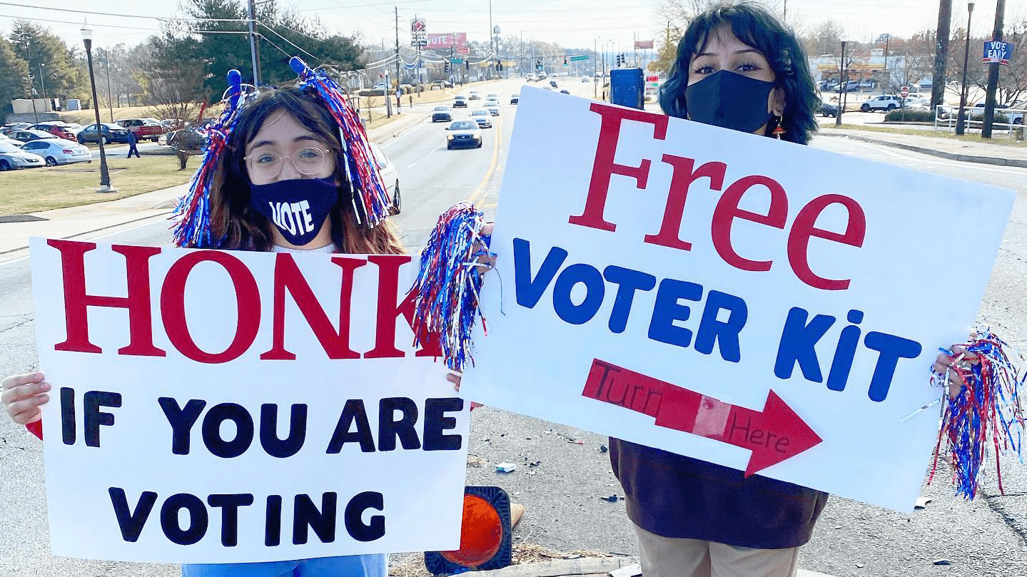 Volunteers with signs that read "honk if you are voting" and "free voter kit."