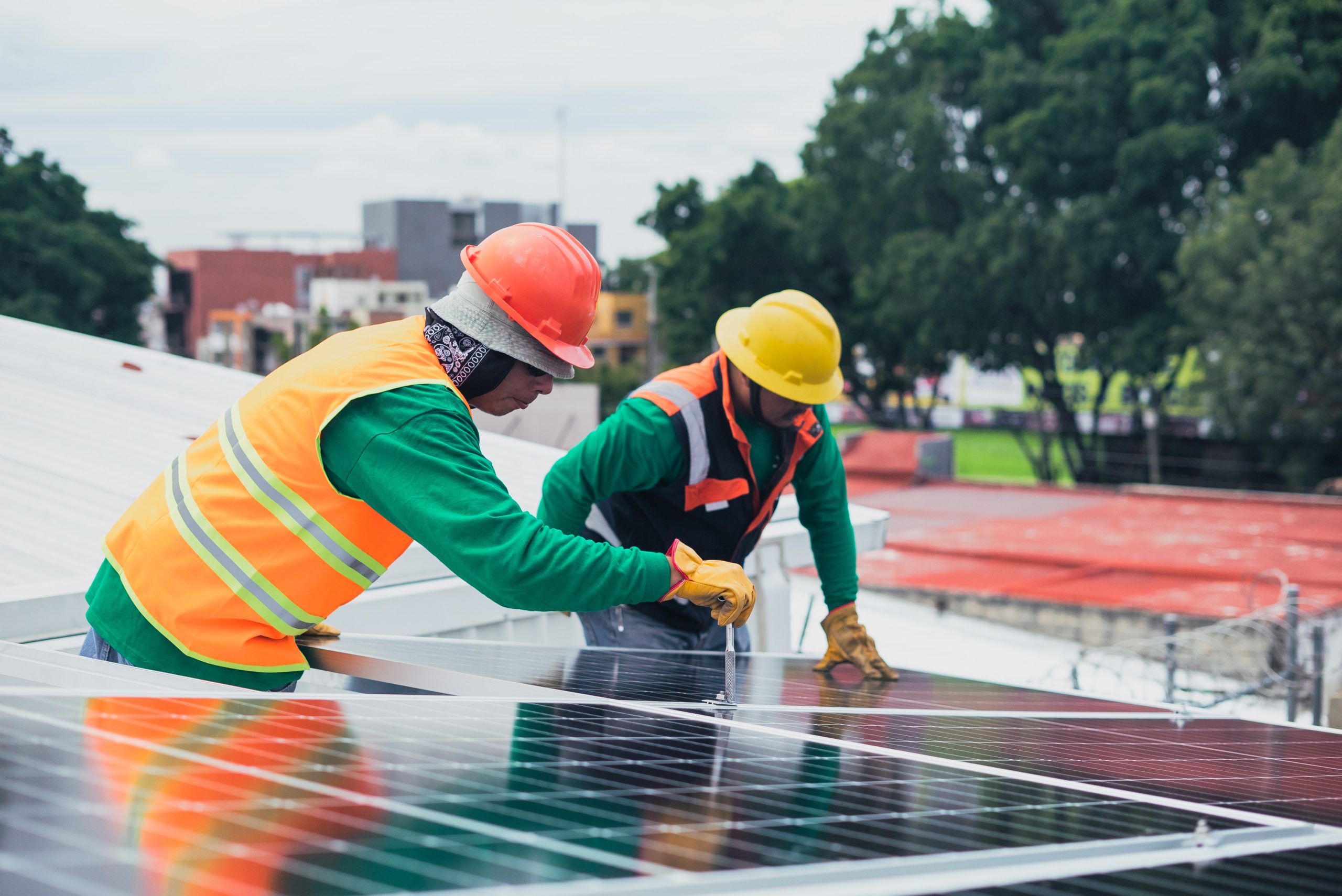 Two construction workers in hard hats lean over a solar panel. One workers is holding a tool.