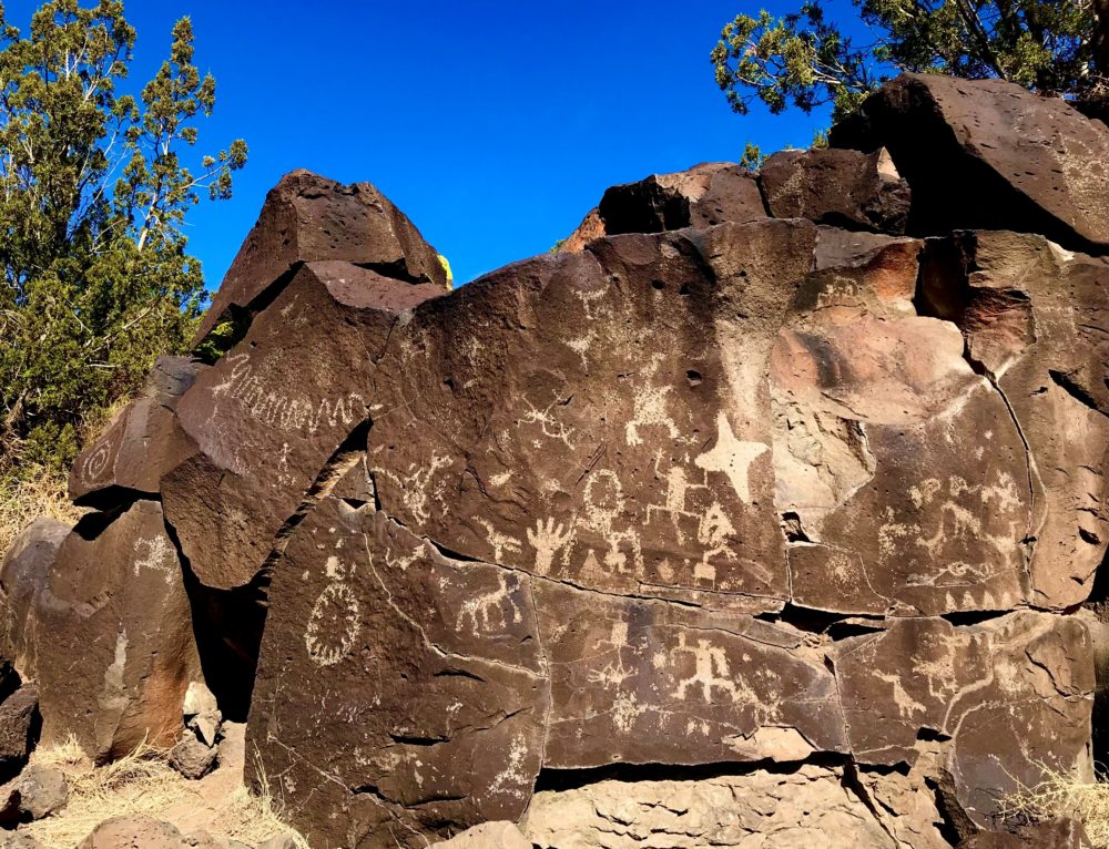 Large, tan and brown boulders stand against a blue sky. There are images carved onto the face of the boulders.