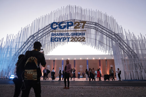 People stand and mingle outside of a building. There are others taking photos in front of a large sign that reads, "COP27 Sharm El-Sheikh Egypt 2022".