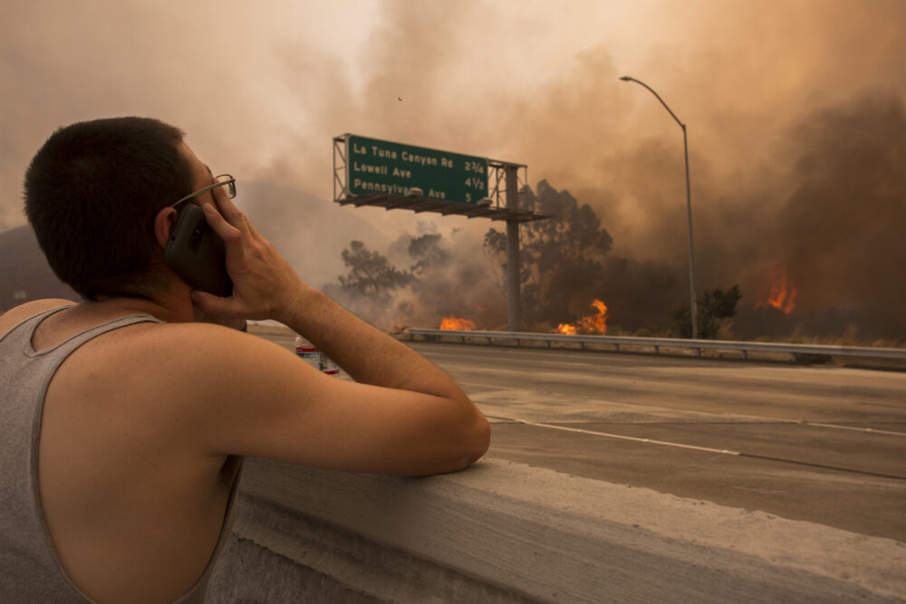 A person holding a phone to their ear watches a fire burning across a highway. the fire is large and is releasing large amounts of smoke.