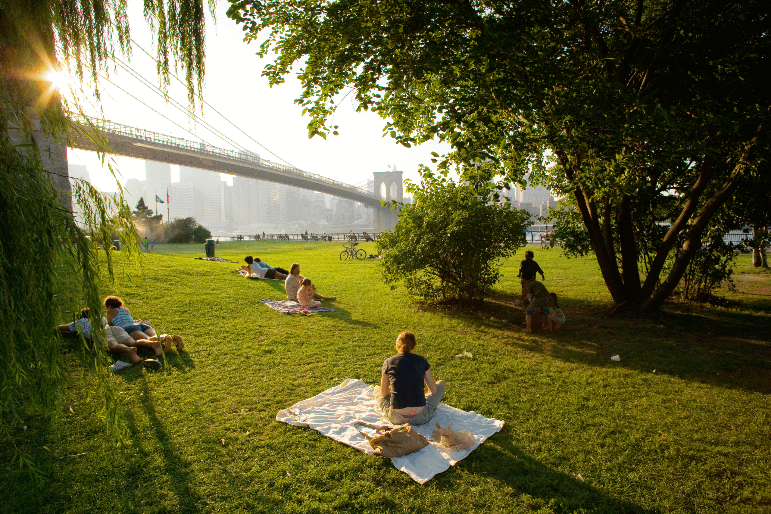Several people and groups of people sit on blankets in a grassy field situated under a bridge.