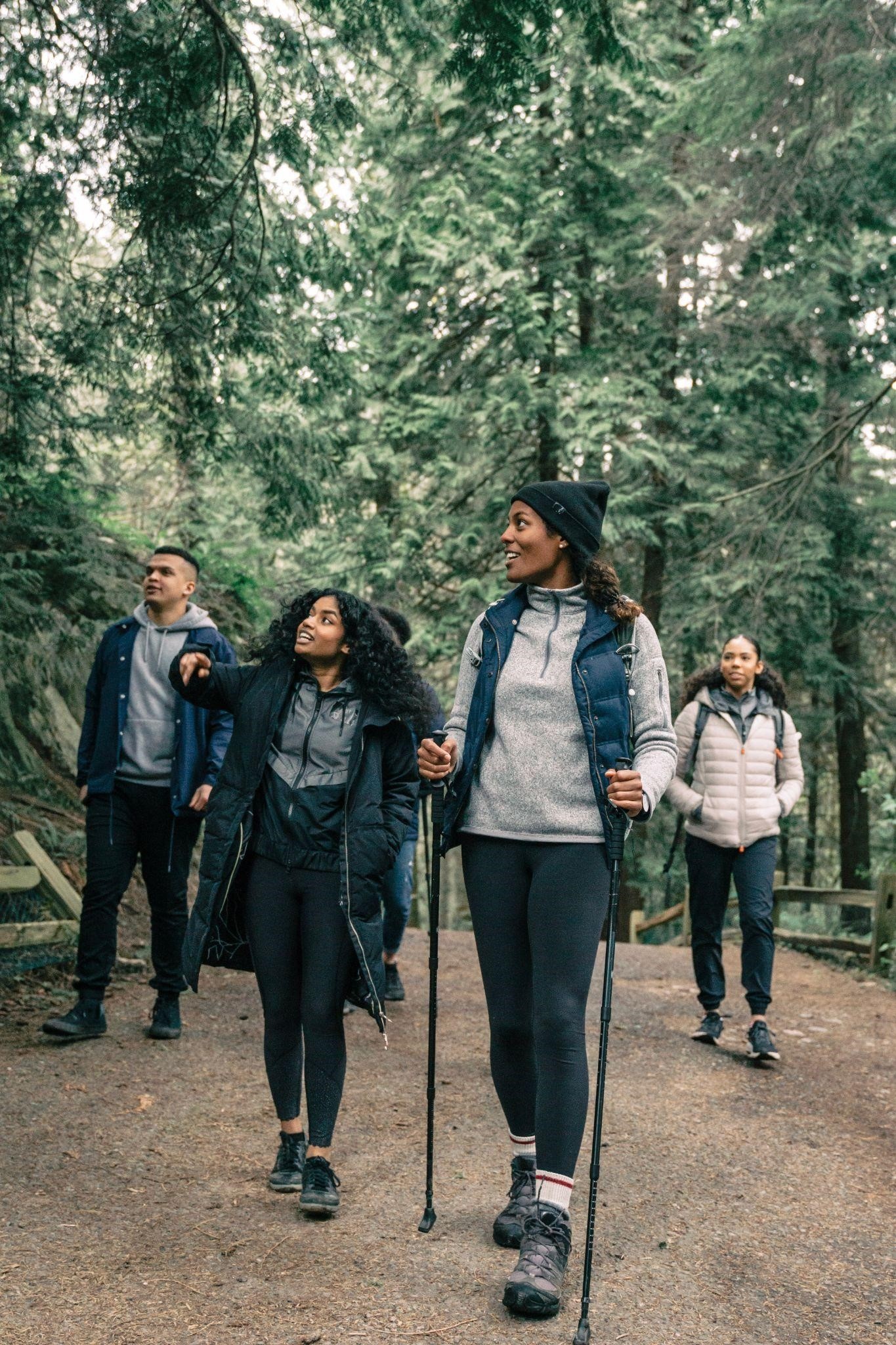 A group of four people are talking and hiking in the woods. One of them is holding two walking sticks.