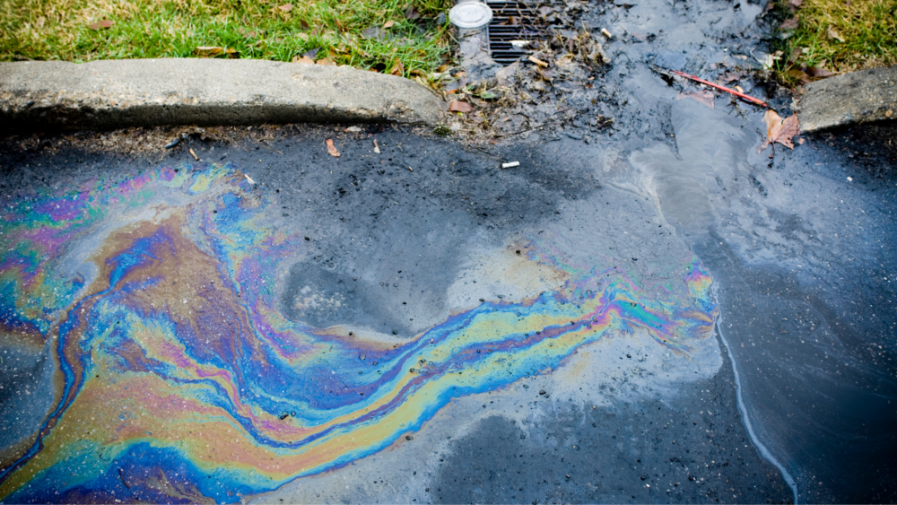A shiny, colorful substance (oil) is flowing towards a metal drainage grate on the street.