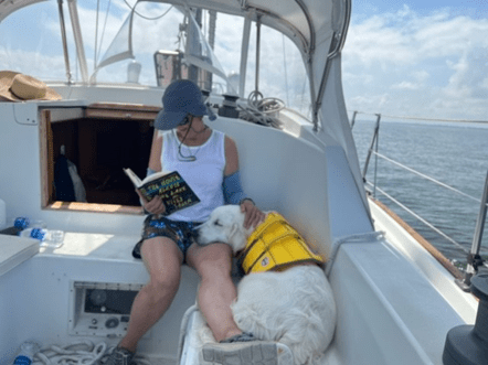 A person on a boat reads a book. There is a dog sitting next to them.