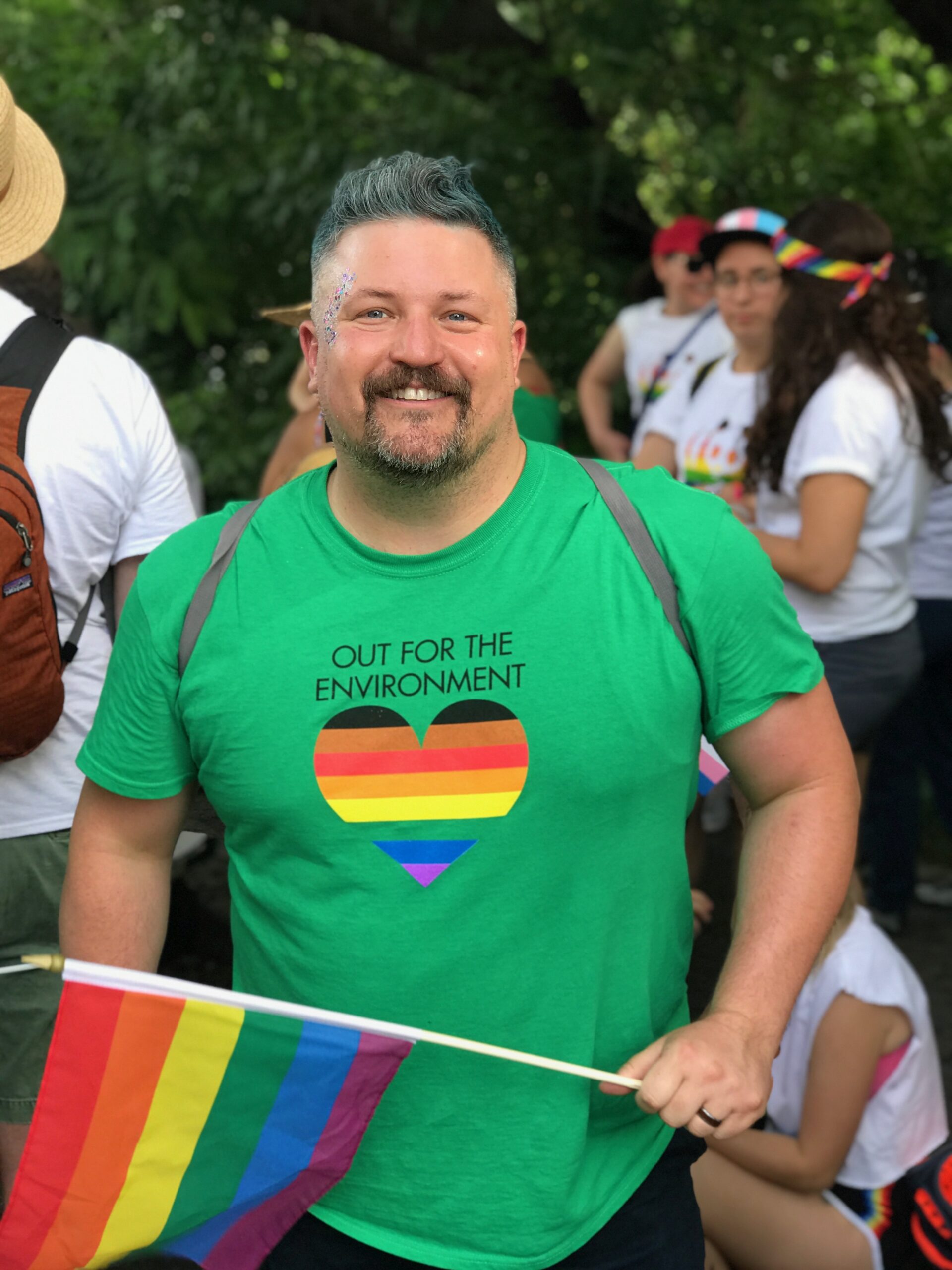 A person wearing a green t-shirt that reads, "OUT FOR THE ENVIRONMENT" is holding a rainbow flag.