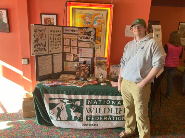 A person in a baseball cap stands beside a table with an older National Wildlife Federation logo on it.