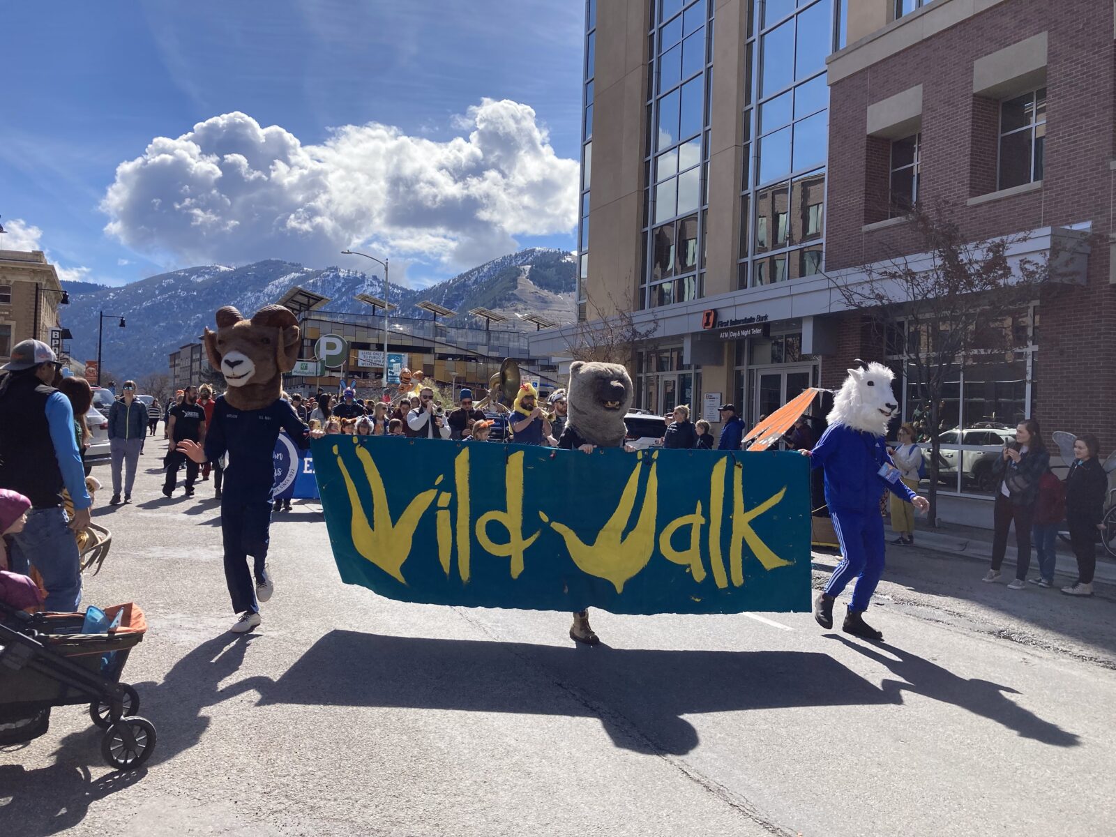 People in costumes and march-goers walk down a street holding a large banner reading, "Wild Walk".