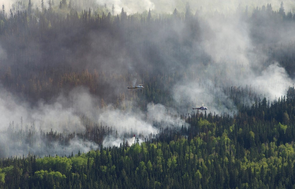 Two helicopters can be seen in the distance flying through thick smoke, coming from a forest below.
