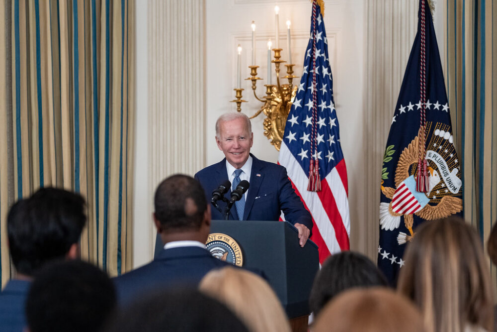 President Joe Biden stands behind a podium. There are reporters in the crowd before him.