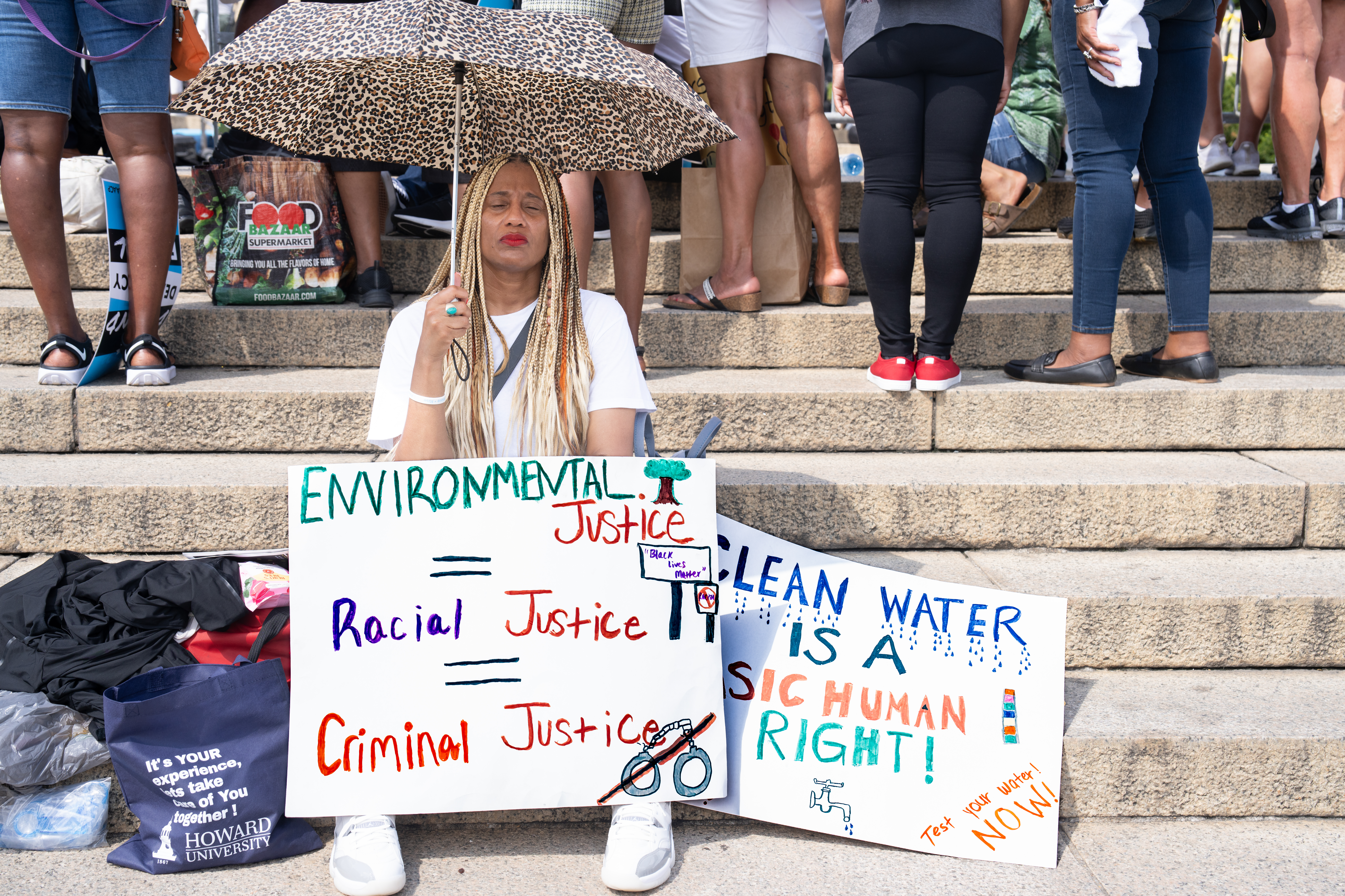 A Black person with box braids sits on the ground with signs in front of them advocating for environmental justice and clean water.