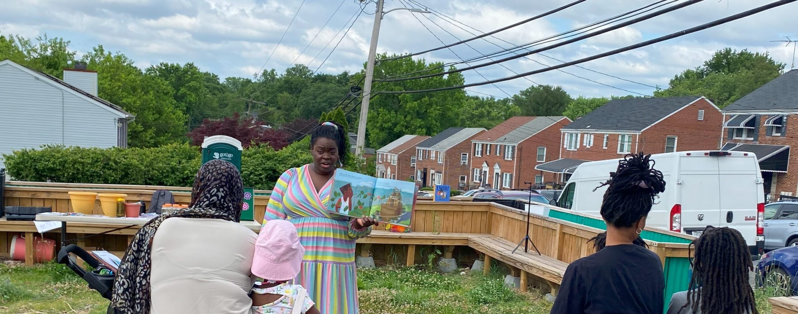 A person stands outside holding a picture book open in front of a small group of adults and children.
