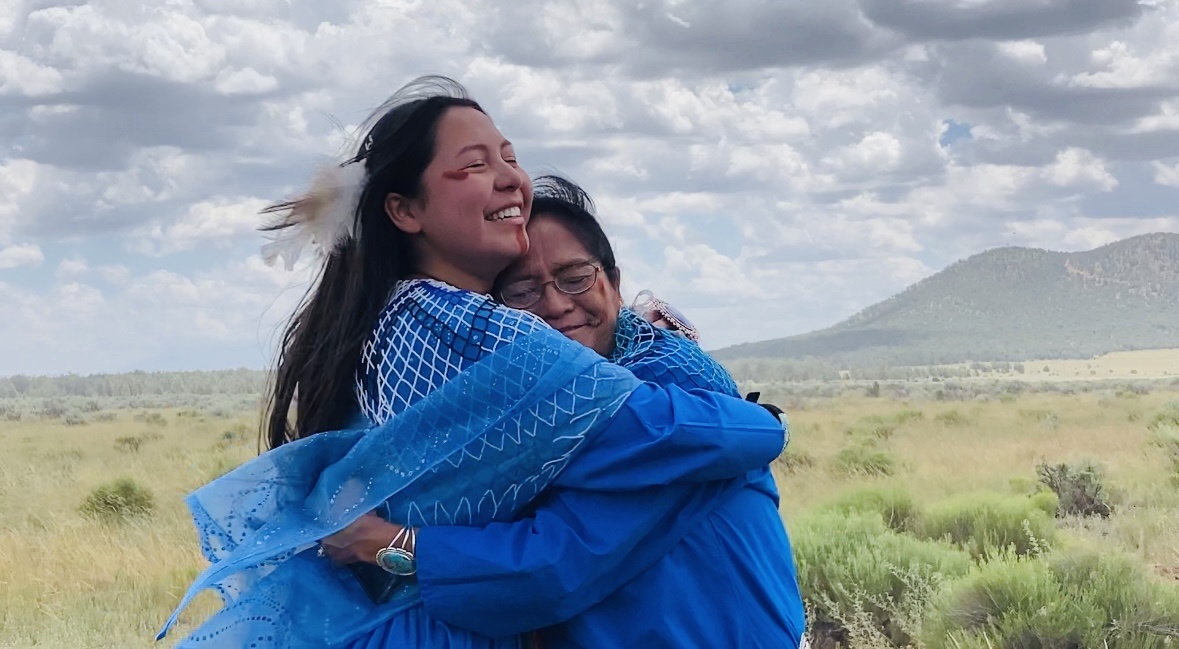 Two people share a heartfelt hug against a prairie landscape.