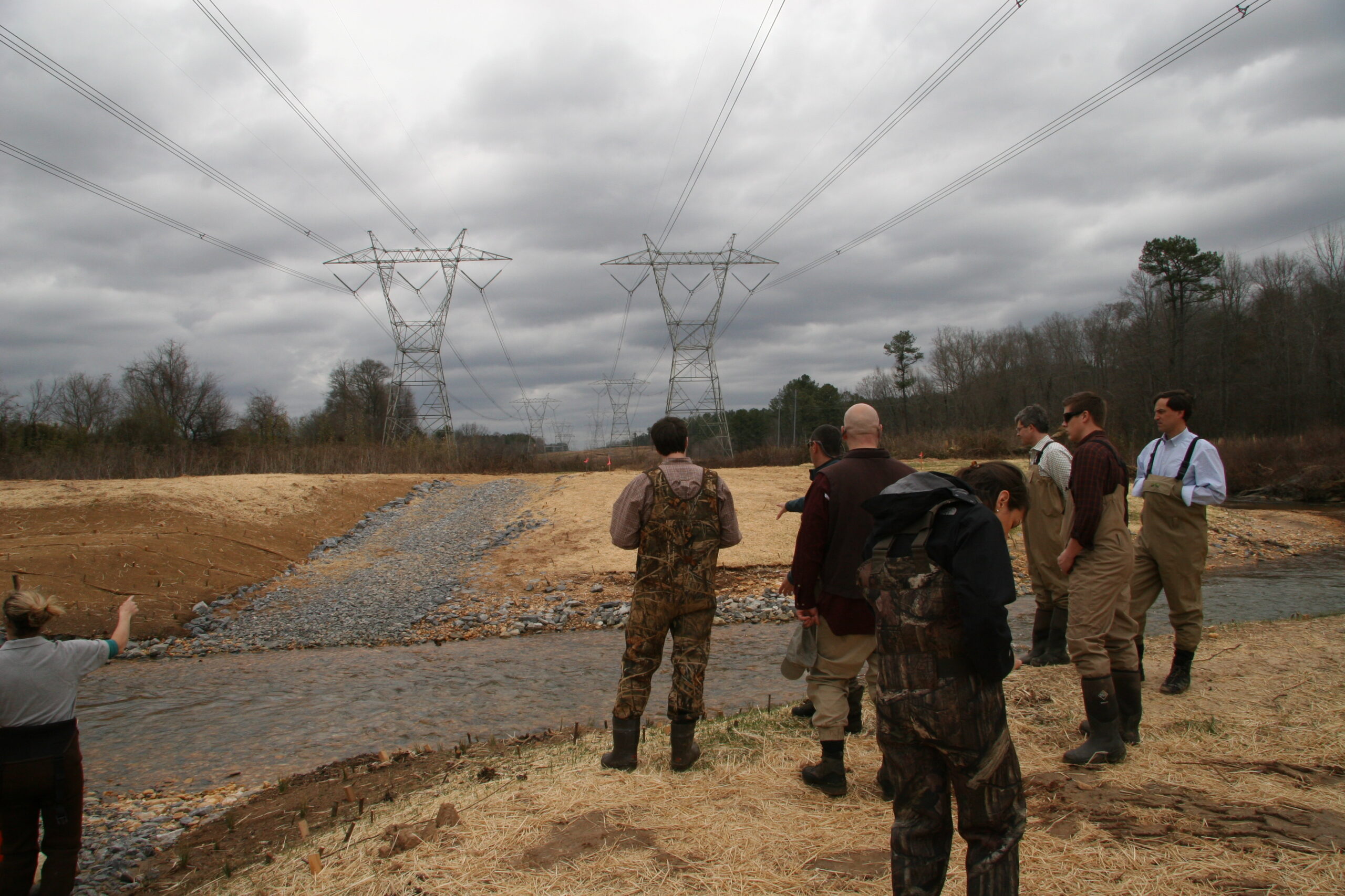 A group of people stand in front of a narrow river looking out onto tall transmission lines. Some are wearing camo gear and boots.
