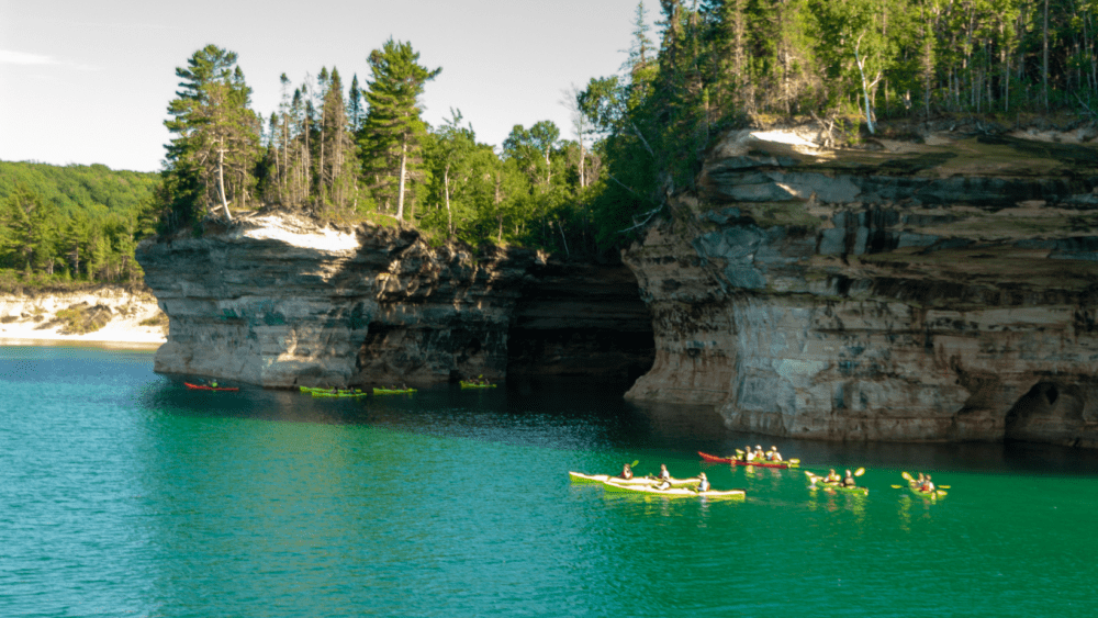Groups of people kayak beneath a cliff.