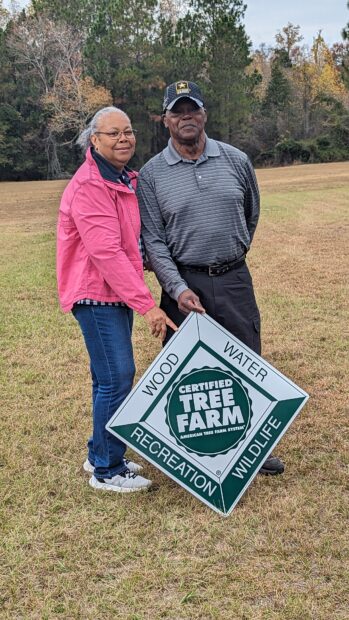Two people hold a sign that says, "Certified Tree Farm - Wood, Water, Recreation, Wildlife".