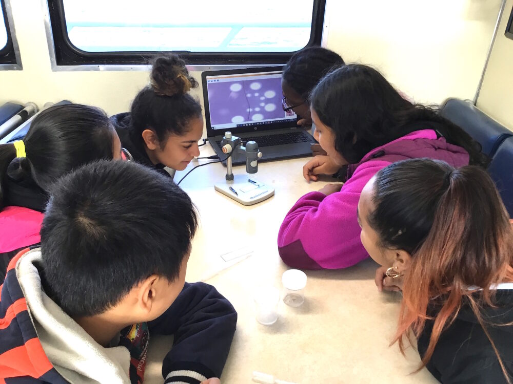 A group of students huddle around a microscope.