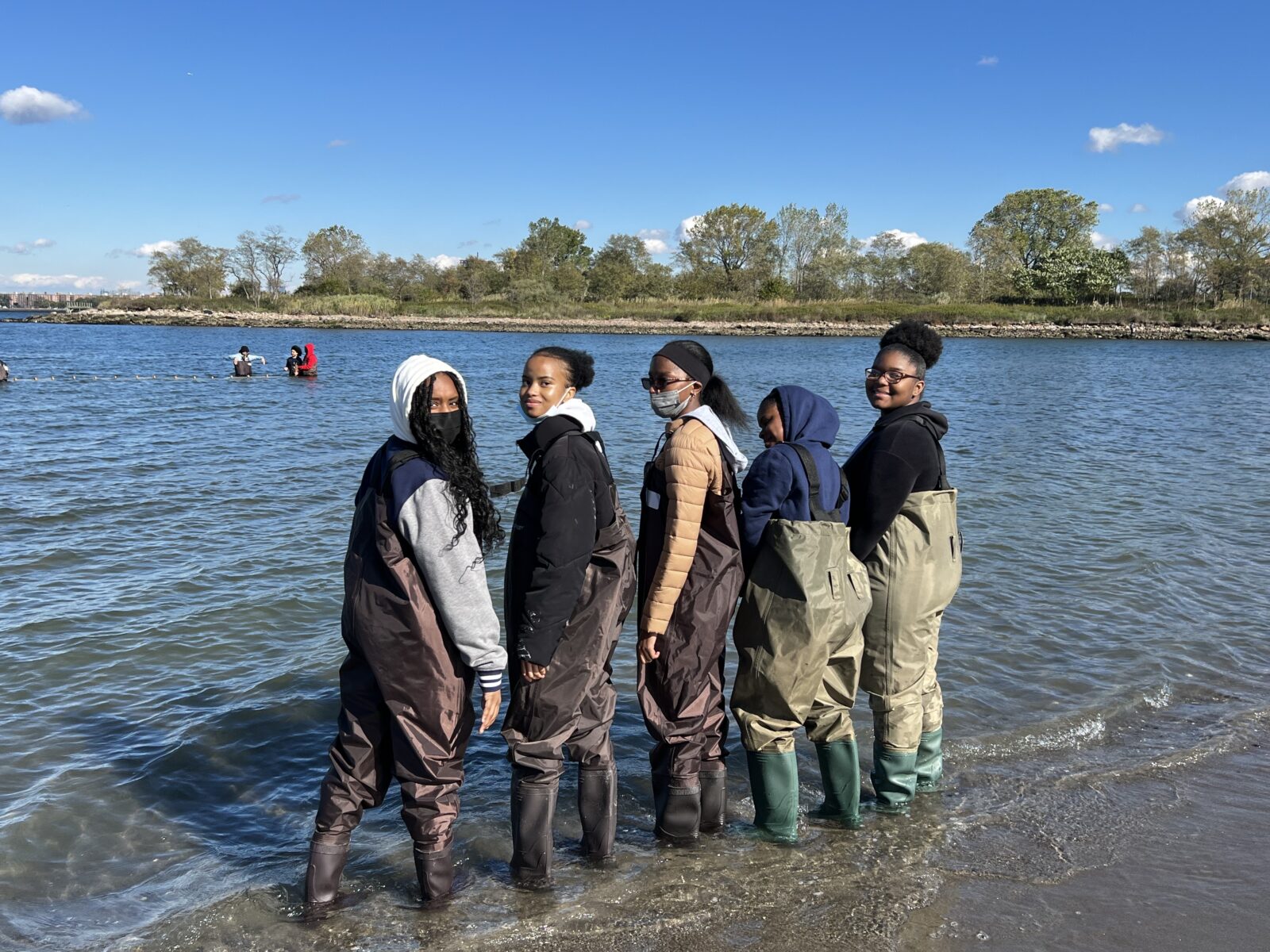 Five young people wearing waders stand on a shore looking back at the camera.