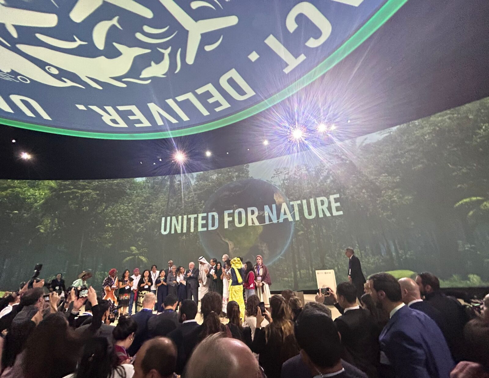 A group of people stand on a stage in front of an audience. A large screen behind those onstage reads, "UNITED FOR NATURE".