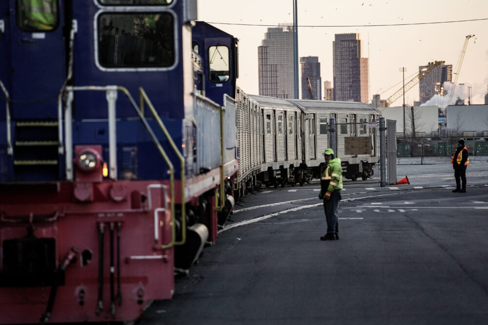 A person in reflective gear stands beside a train. Buildings can be seen in the distance.