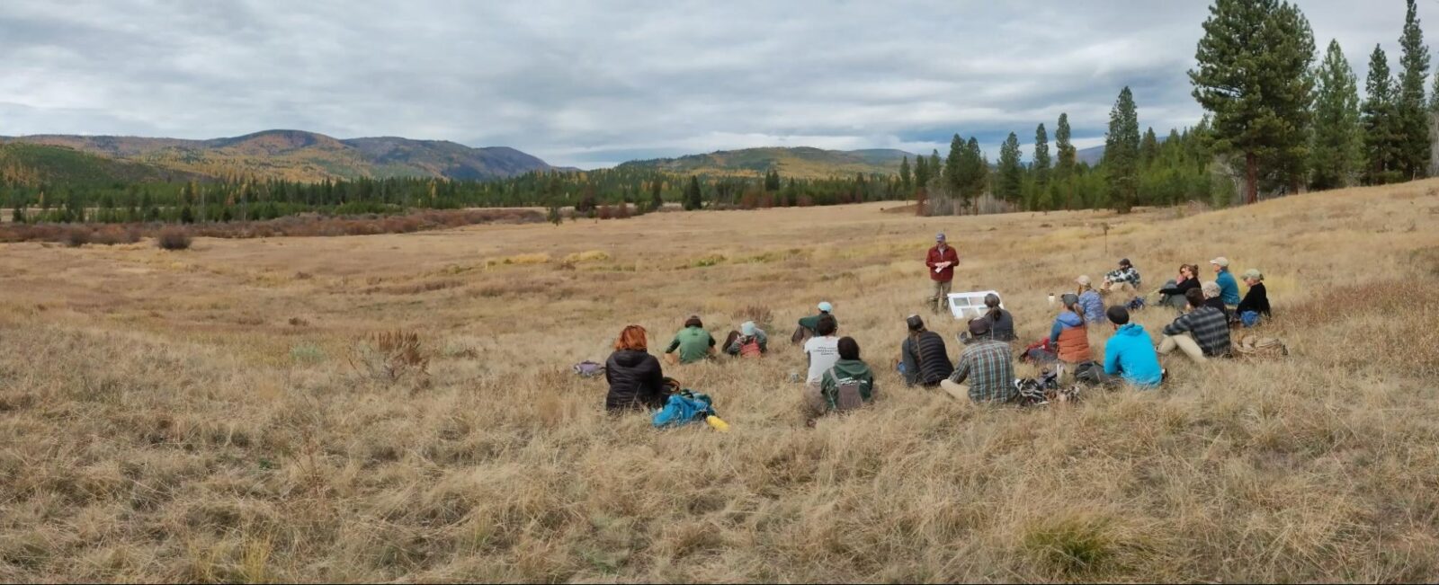 A group of people sit in a large field, mountains are seen in the distance.