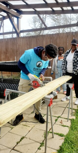 A young volunteer wearing a blue "Earth Tomorrow" t-shirt uses a circular saw to divide a plank of wood.