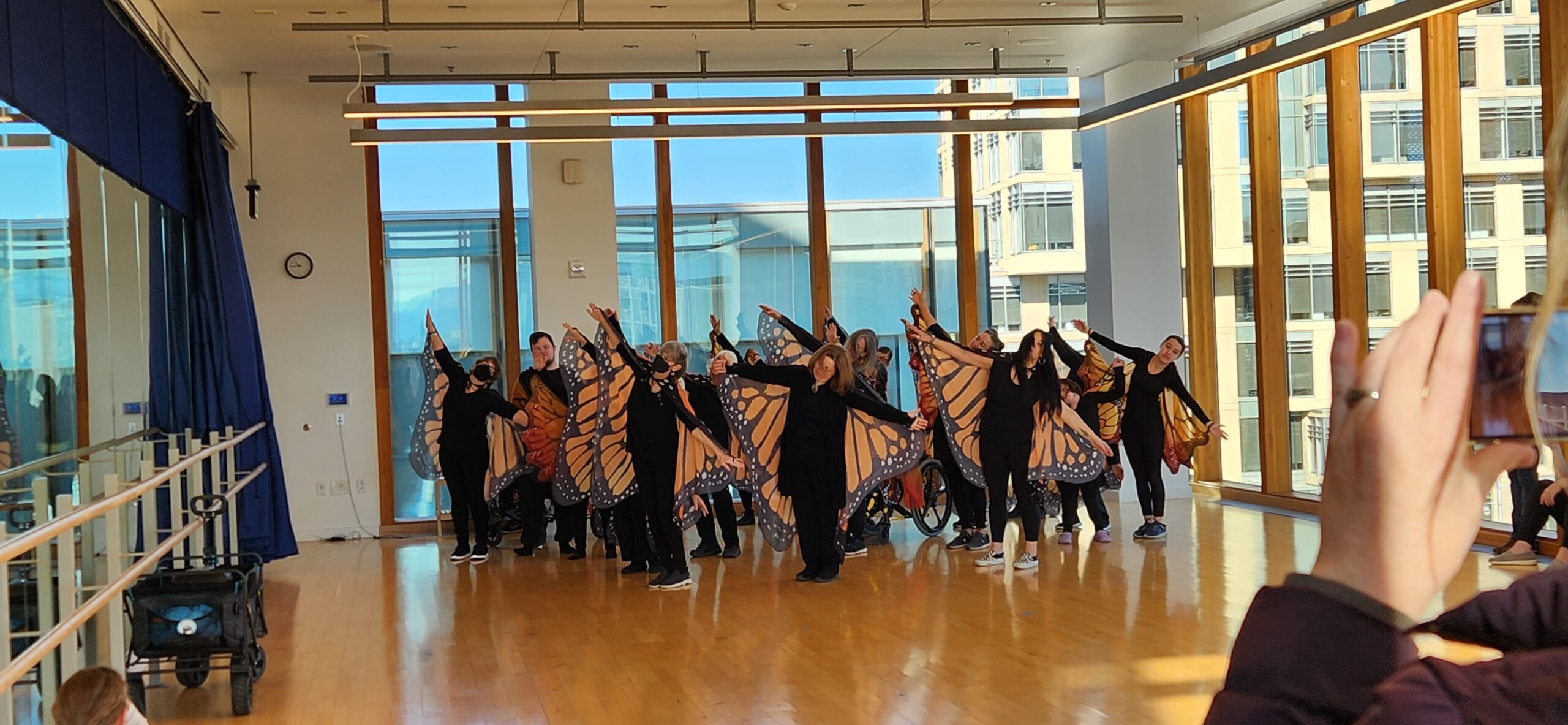 A group of people stand in formation in a dance studio. They are wearing all black clothing and each person has a pair of fabric monarch wings attached to them.