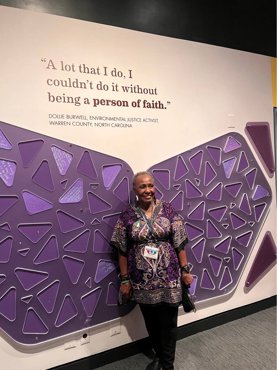 A Black woman (Dollie Burwell) poses for a picture against a wall featuring a quote by her that reads, "A lot that I do, I couldn't do without being a person of faith."