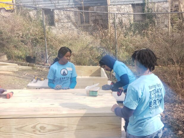Young volunteers wearing blue "Earth Tomorrow" t-shirts work on a wooden garden box.