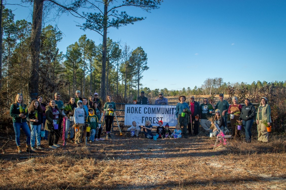 A large group of people gather for a photo around a banner that reads, "Hoke Community Forest".