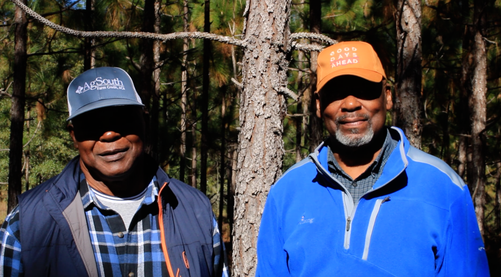 Two Black men wearing caps stand in a forest and pose for a photo.