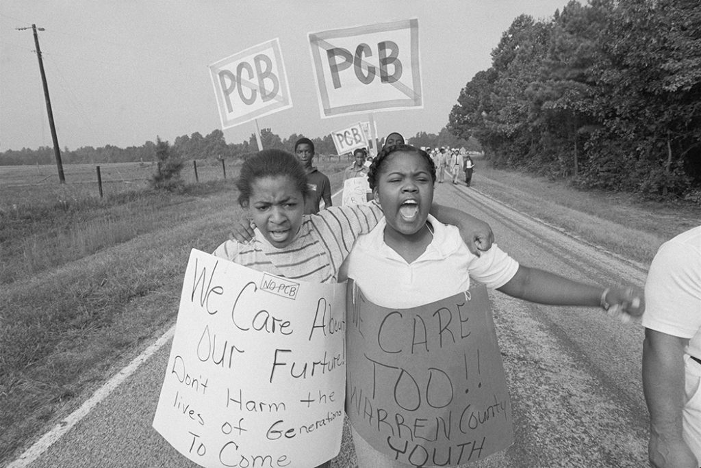 Two Black people wearing posters on their bodies are seen marching down a road with other protesters.