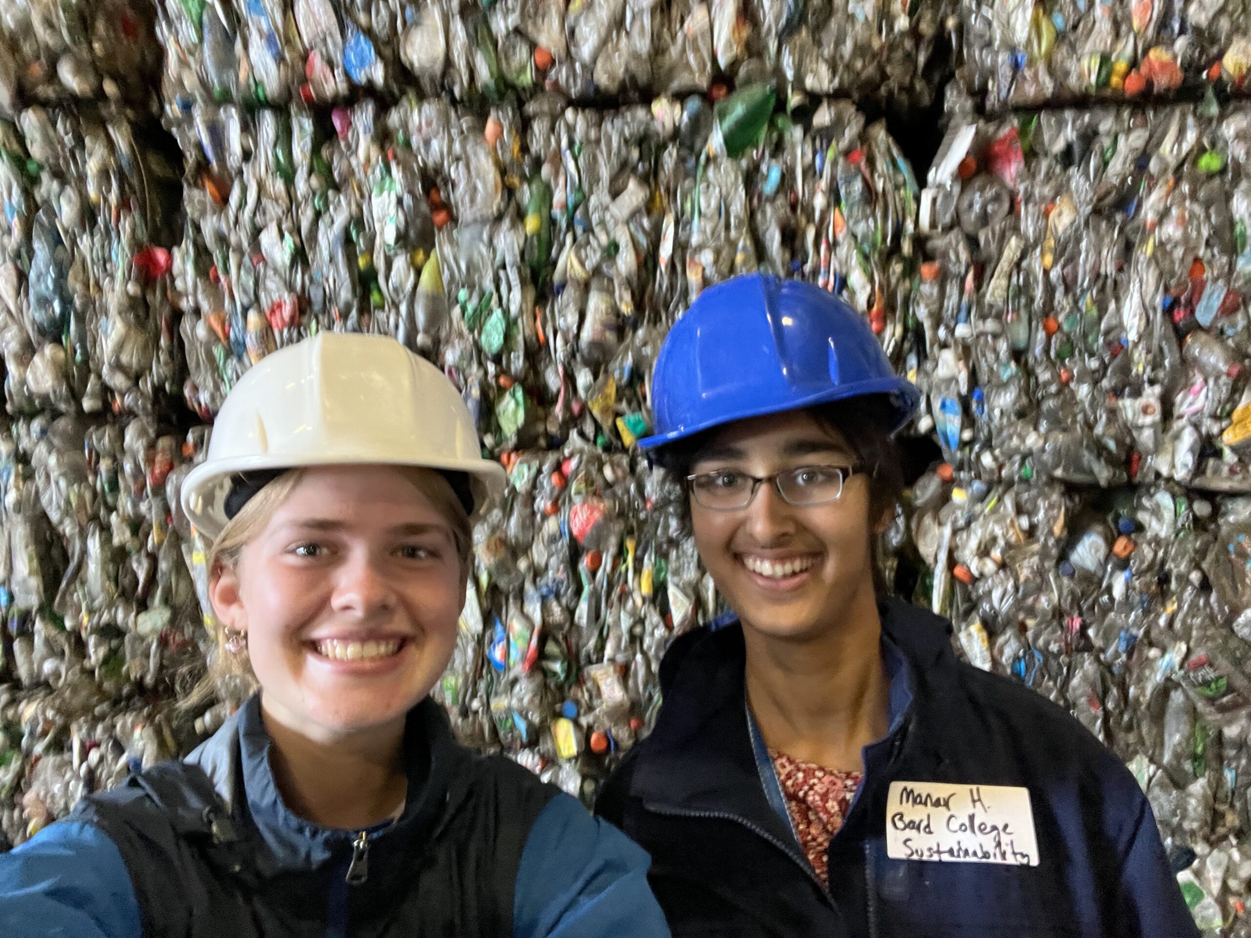 Two young people wearing hard hats smile for a selfie in front of compressed blocks of recycled materials.