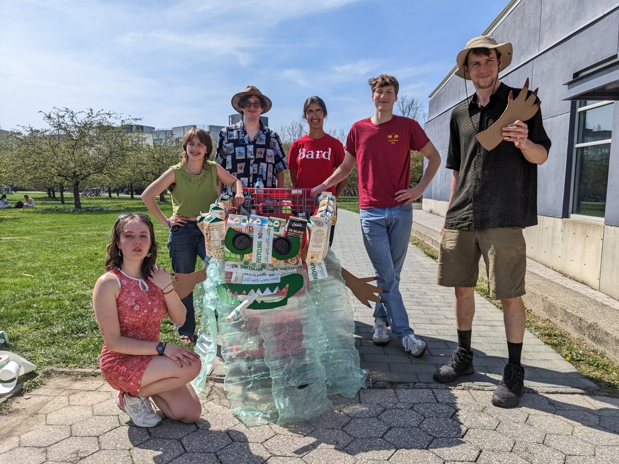 Six young people pose outside with a piece of art made from recycled materials. The art piece has eyes and pointy teeth.