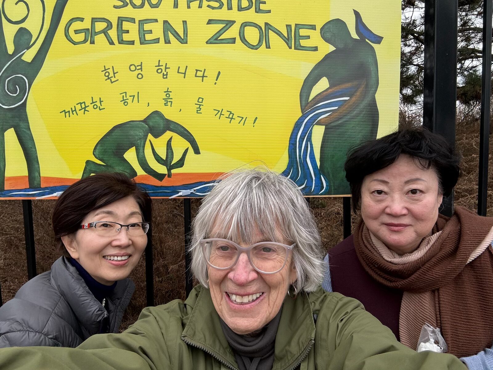 Three people pose for a selfie in front of a banner that reads, "Green Zone". There is more text in another language on the banner.