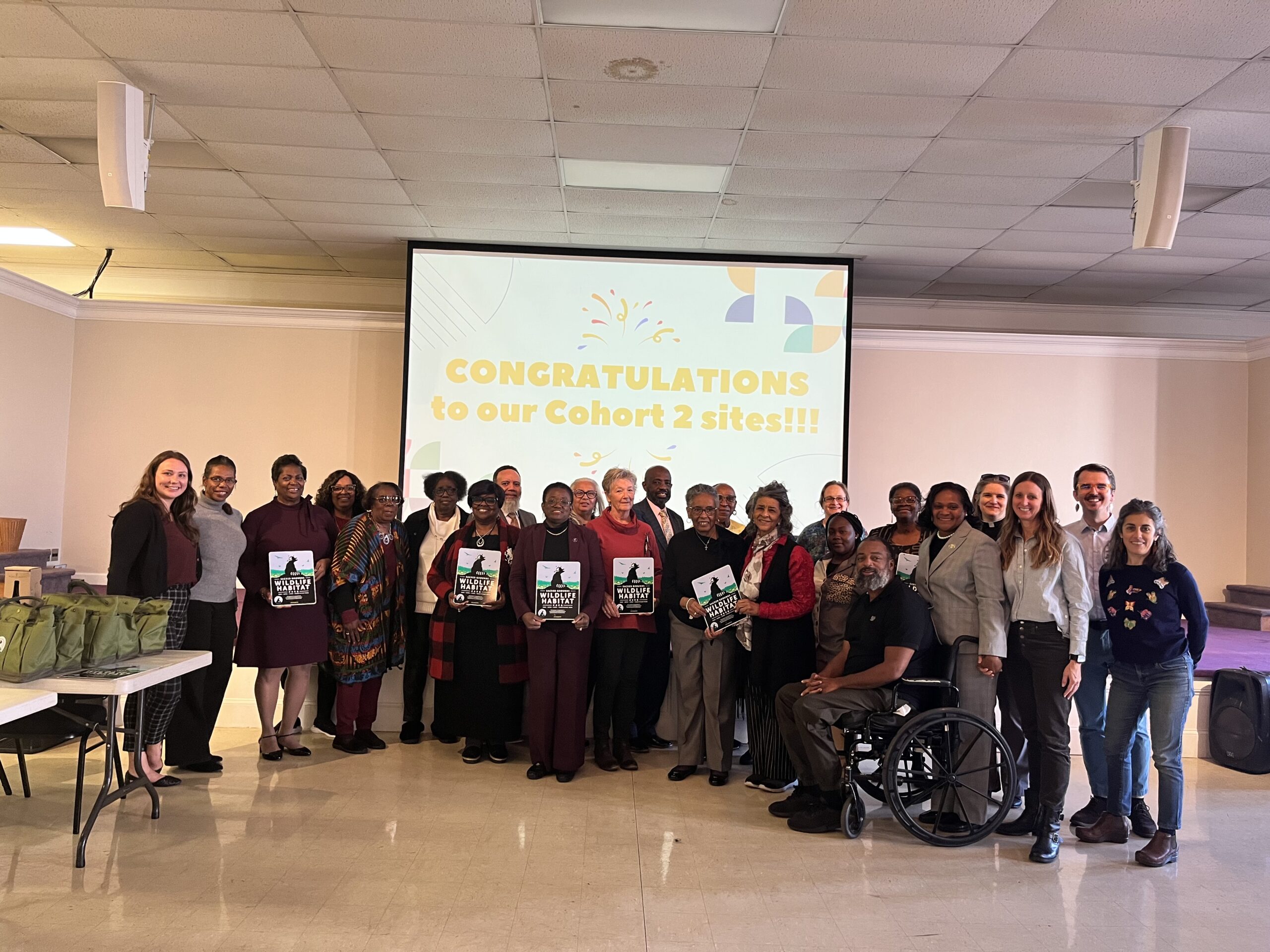 A large group of people pose for a photo indoors in front of a projector screen that reads, "Congratulations to our cohort 2 sites!!!".