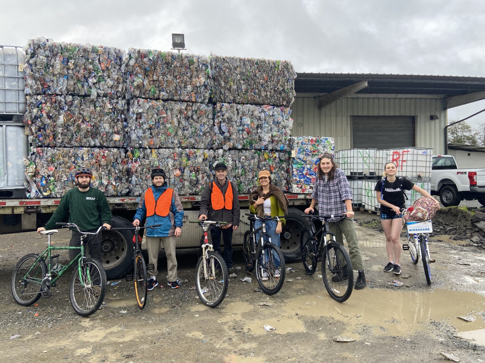 Six young people pose with bicycles in front of a truck bed loaded with compressed blocks of recycled materials.