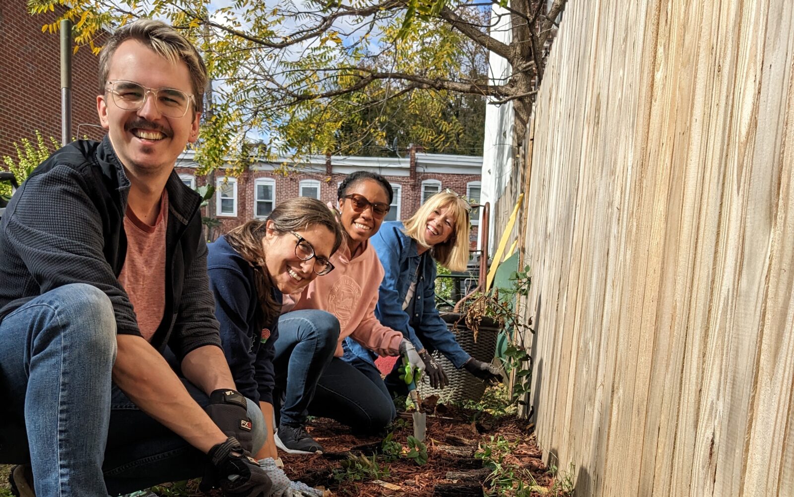 Four people squat down to the ground and plant seedlings in a garden bed. They are facing the camera and smiling.
