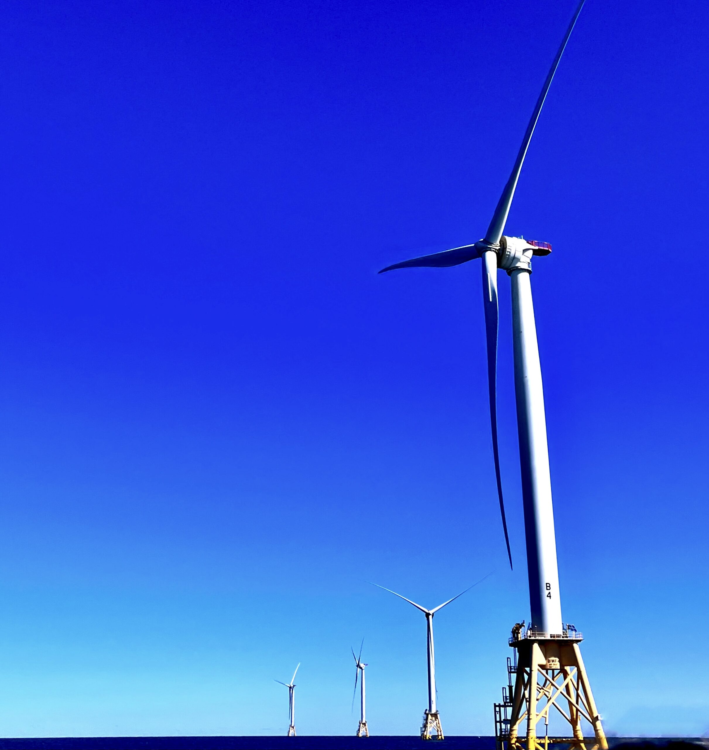 Severl wind turbines stemming from the water stand tall against a bright blue sky.
