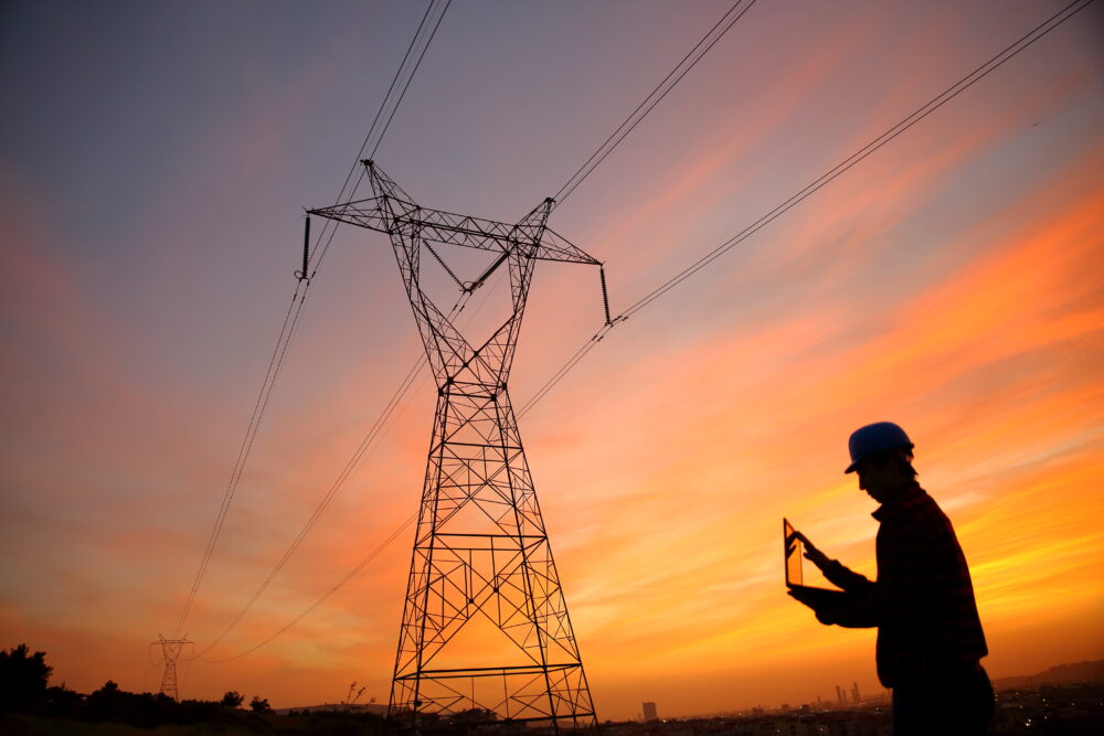 Against a setting sun, a worker wearing a hardhat is holding a laptop. A transmission tower can be seen.