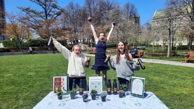 Three people pose outside for a photo in front of a blanket with plants and reusable water bottles on display.