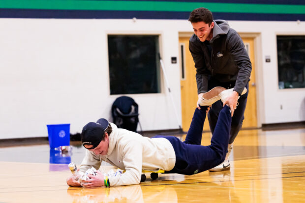 Two people are in a gymnasium playing a game. One person holds the other by their ankles.