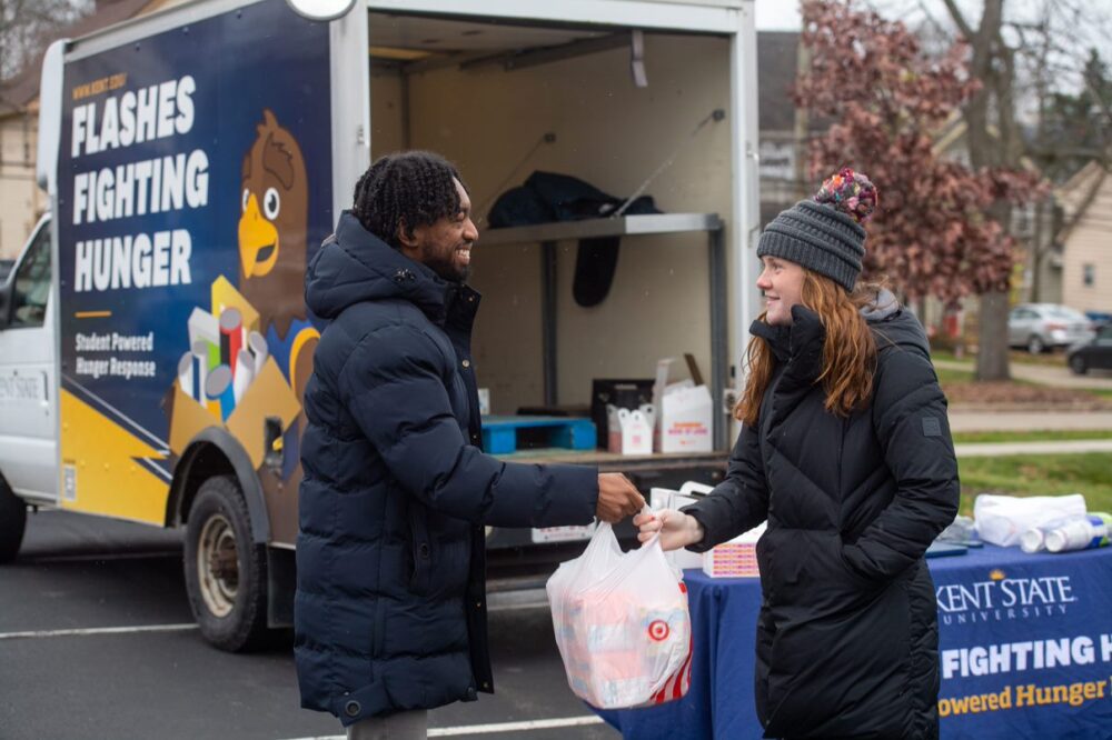 Two people in winter gear are outside in front of a truck and booth. The truck reads, "Flashes Fighting Hunger". One person hands a bag to the other.