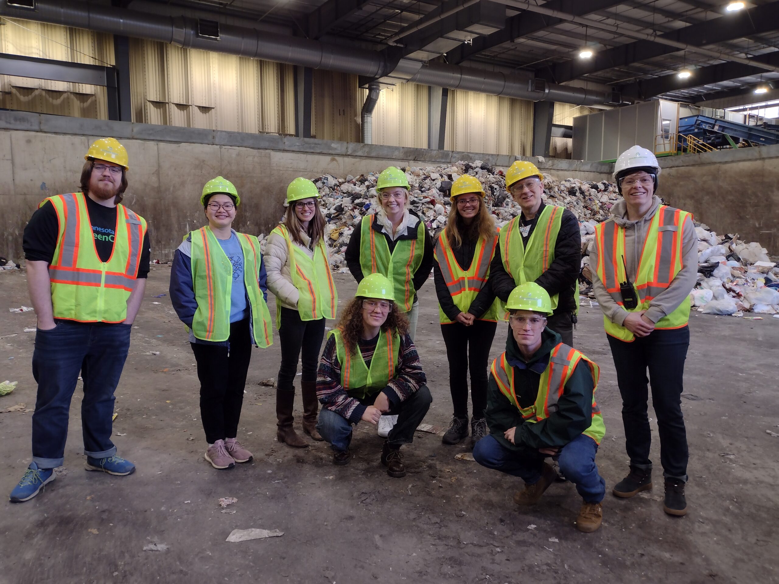 A group of people wearing reflective vests and hard hats pose for a photo at a waste management facility.