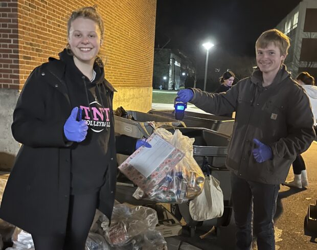 Two people stand outside near a few trash bins. They are using a gravity scale to weigh bags of waste.