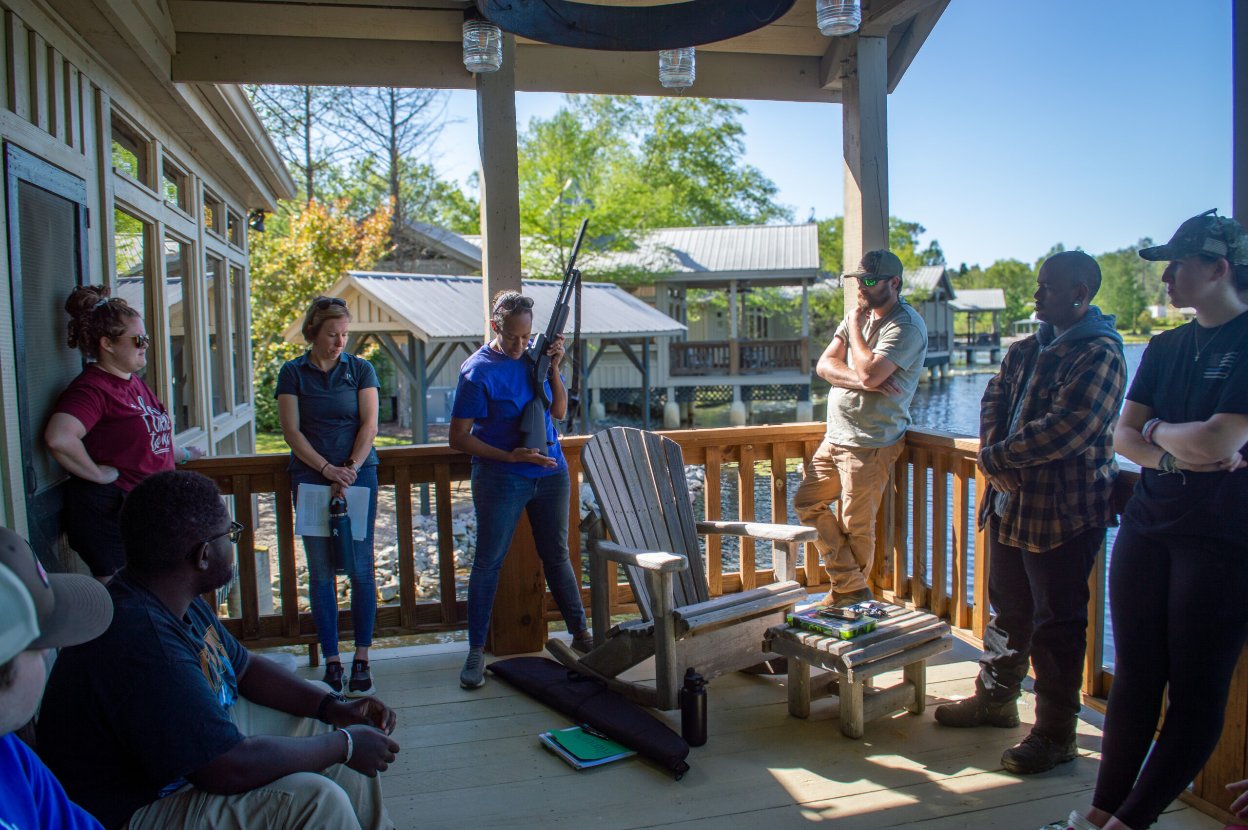 A group of people stand around on a deck overlooking water. One of them is holding a gun and demonstrating how to use it.