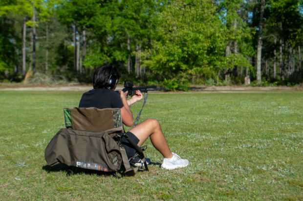 A person sitting in a low chair and wearing ear coverings holds a gun and aims it straight forward.