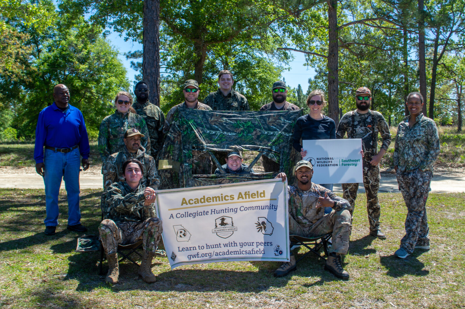 A group of people pose for an outdoor photo. Many of them are wearing camouflage. Two of them are holding a banner that reads, "Academics Afield: A Collegiate Hunting Community".