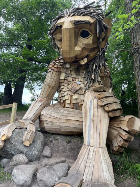 A wooden sculpture depicts a troll with its legs crossed, one eye closed, and tongue stuck out.