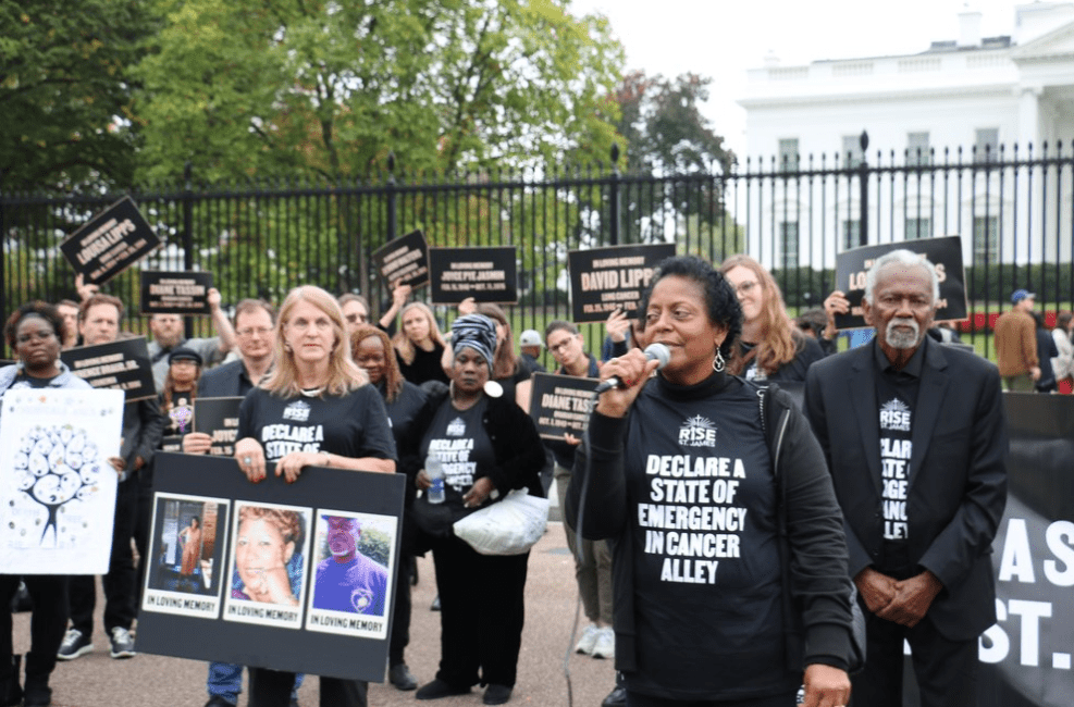 A group of people marching in front of the White House all wear t-shirts that read, "Declare a State of Emergency in Cancer Alley." A person in the front is speaking on a microphone.